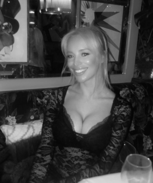 A photograph of Amber Rose sitting in a chair looking at the camera and smiling. She is wearing a black lace top and has a mirror in the background.